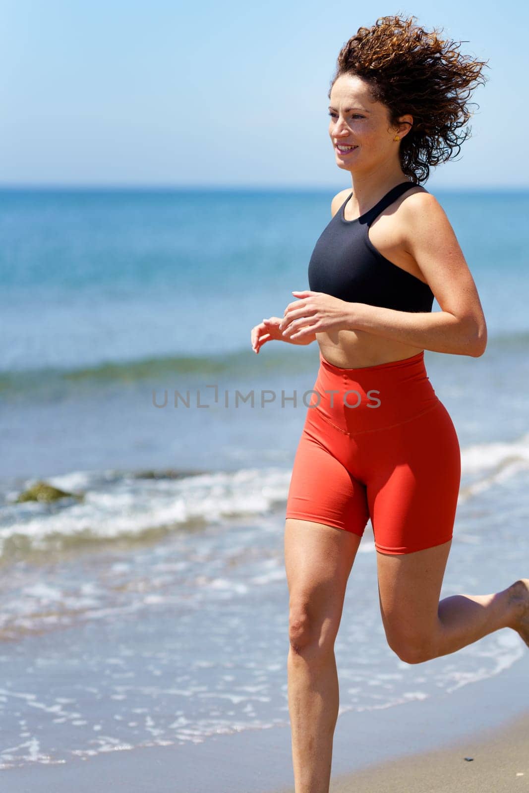 Side view of barefoot young female in sportswear with curly hair smiling and looking away, while jogging in daylight on sandy beach near foamy seawater against blurred blue sky
