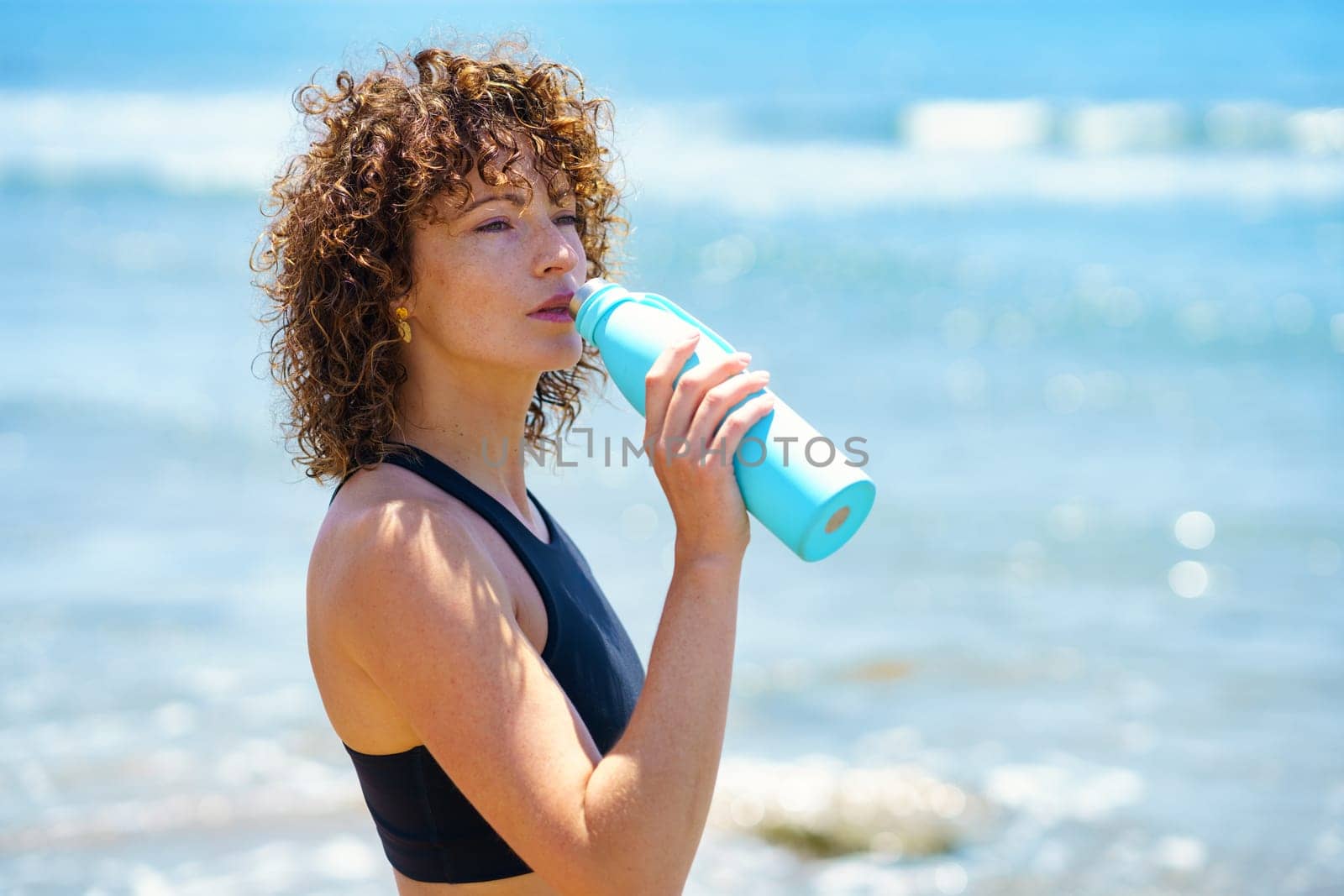 Side view of young female athlete with curly hair standing on seashore and drinking water from blue plastic bottle after workout