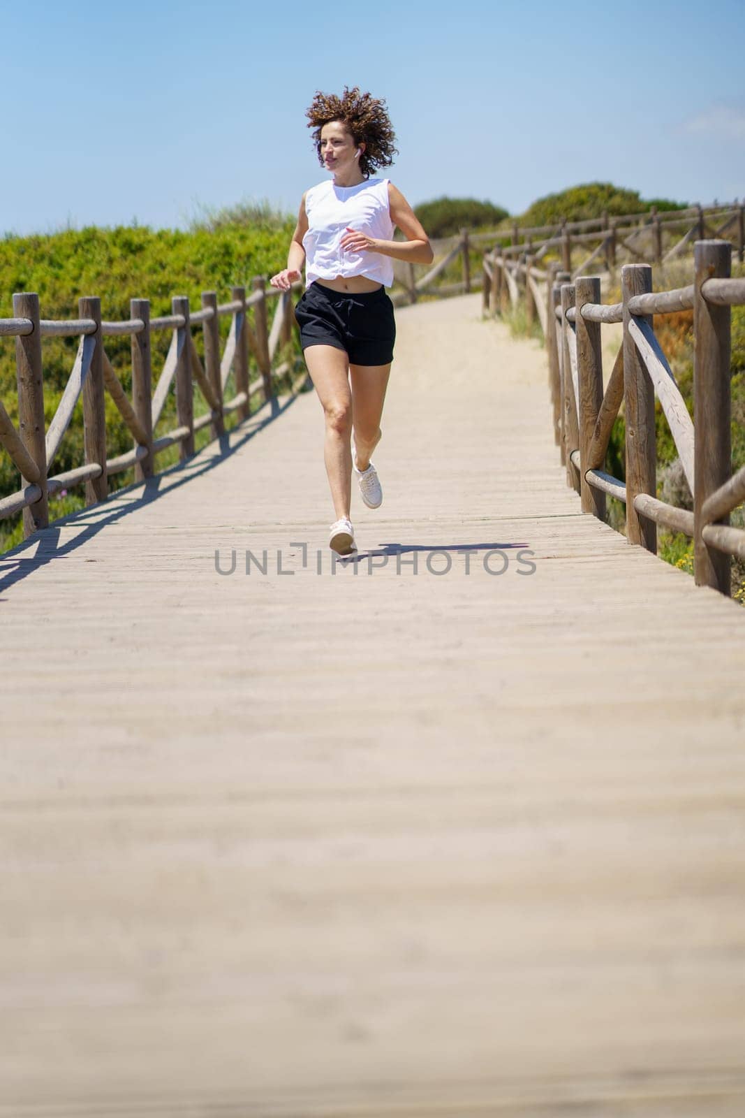 Full body of fit female athlete in sportswear running on wooden path and looking away during sunny summer day