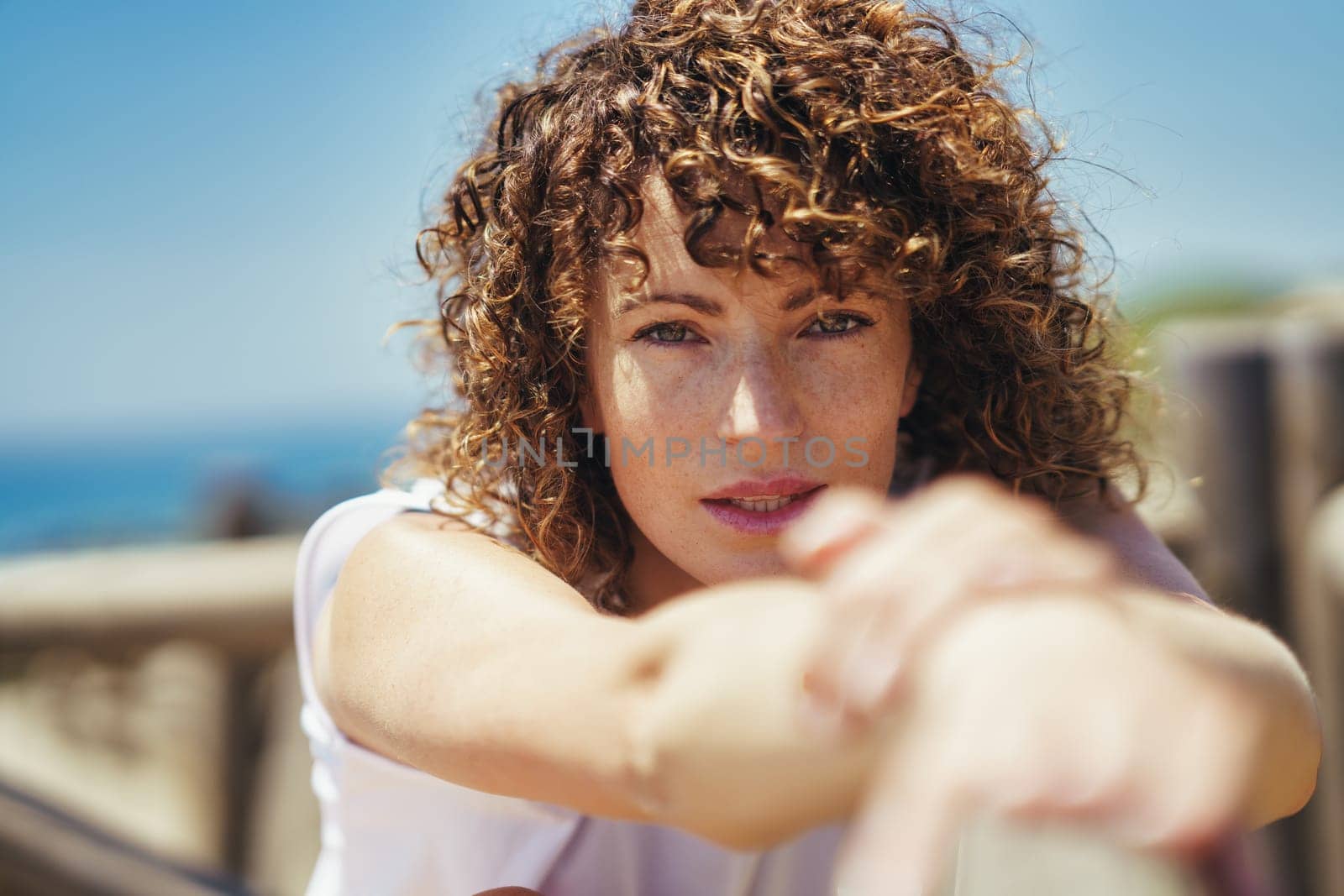 Young curly haired female with freckles and brown wavy hair in white casual clothes stretching arms while leaning on fence against blurred background