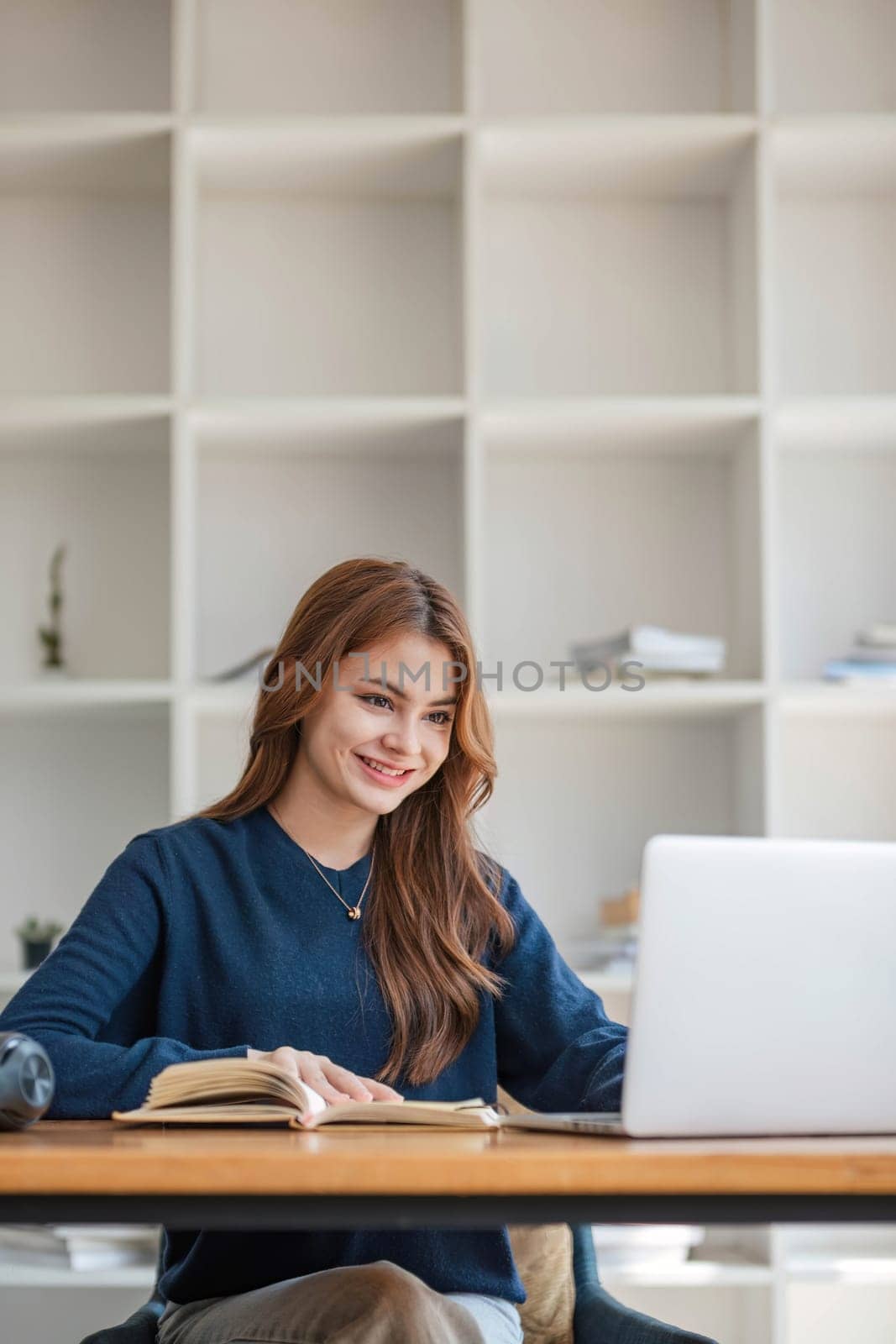 Smiling asian undergraduate teen girl student study in library with laptop books doing online research for coursework, making notes for essay homework assignment, online education e-learning concept by wichayada