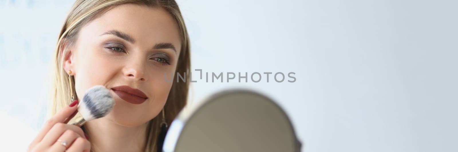 Smiling young woman looks in the mirror and paints herself with blush brushes by kuprevich