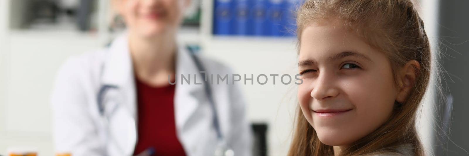 Portrait of child girl winks at camera in doctor office. Child pretending to be sick at pediatrician appointment
