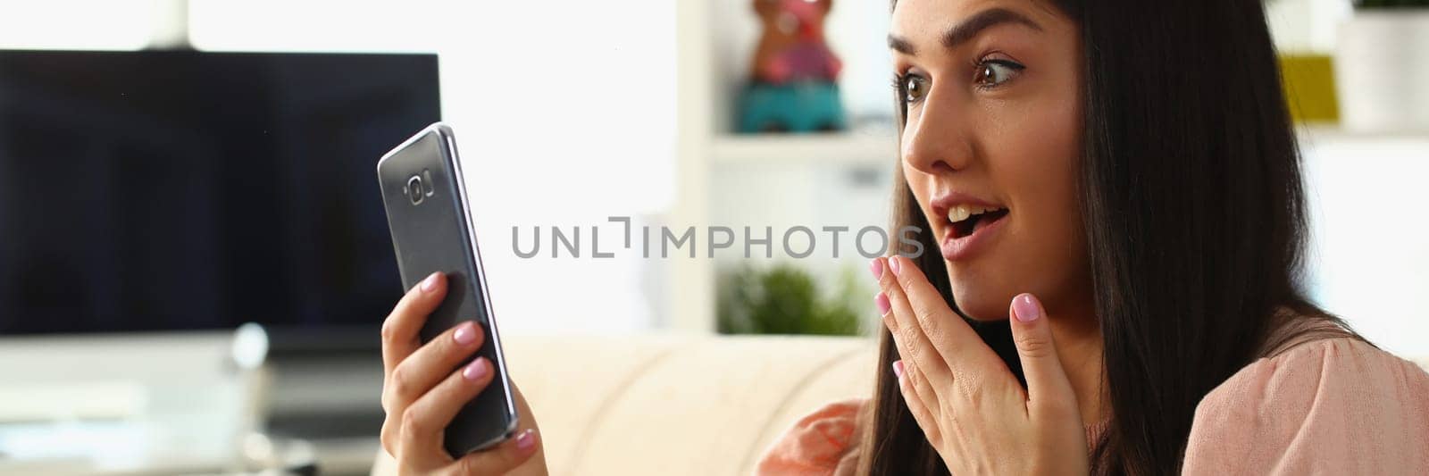 Woman surprised in shock looks at smartphone screen while sitting on couch at home. Emotion joy delight and good news concept