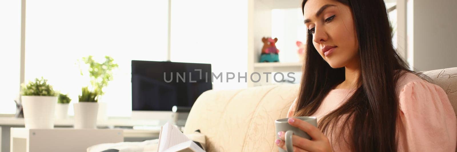 Woman sits on sofa and reads book holding mug of coffee in living room at home by kuprevich