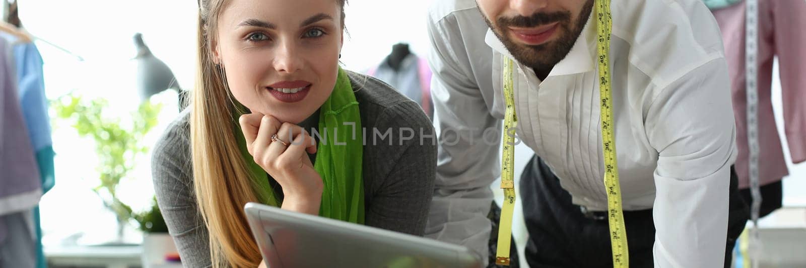 Man and woman designers are discussing new collection of clothes on digital tablet in workshop. Workers of online fashion store in modern warehouse.