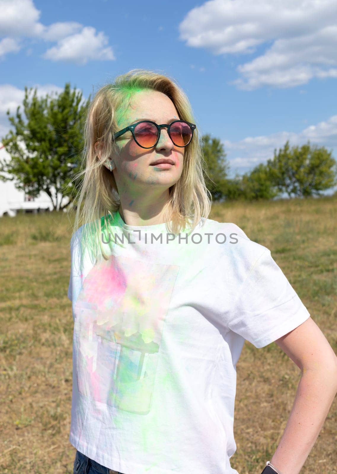 Portrait Pretty Blond Woman With Colored Dye, Powder On Face And Cloth On Holi Colors Festival In Meadow, Sunny Day. Playful Cultural Event With Throwing Bright Neon Powder. Vertical Plane.