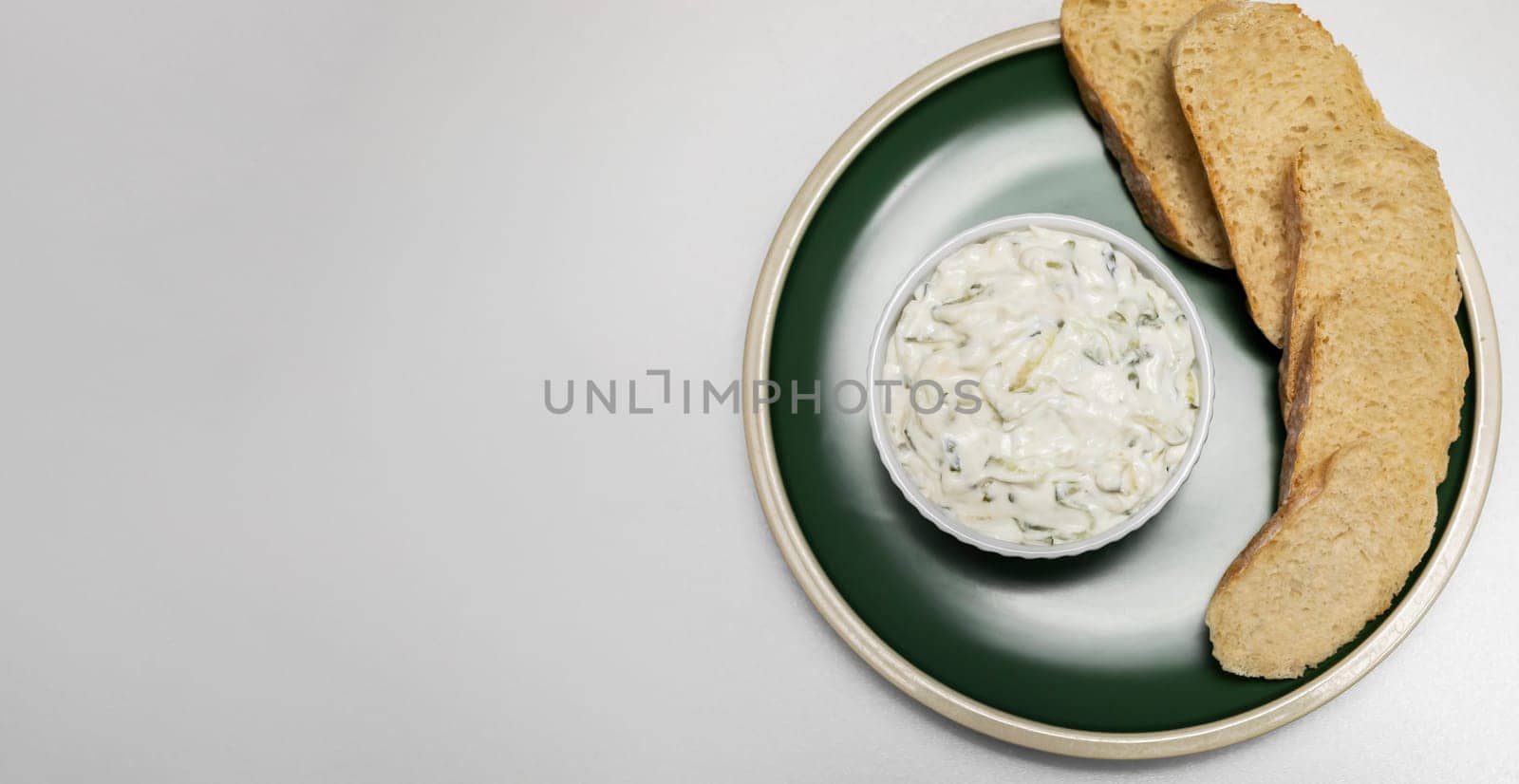 Traditional homemade greek yogurt sauce Tzatziki, tarator in white bowl, made of grated cucumber, sour cream yogurt,olive oil,garlic,herbs on green plate with sliced bread, white background.Copyspace.