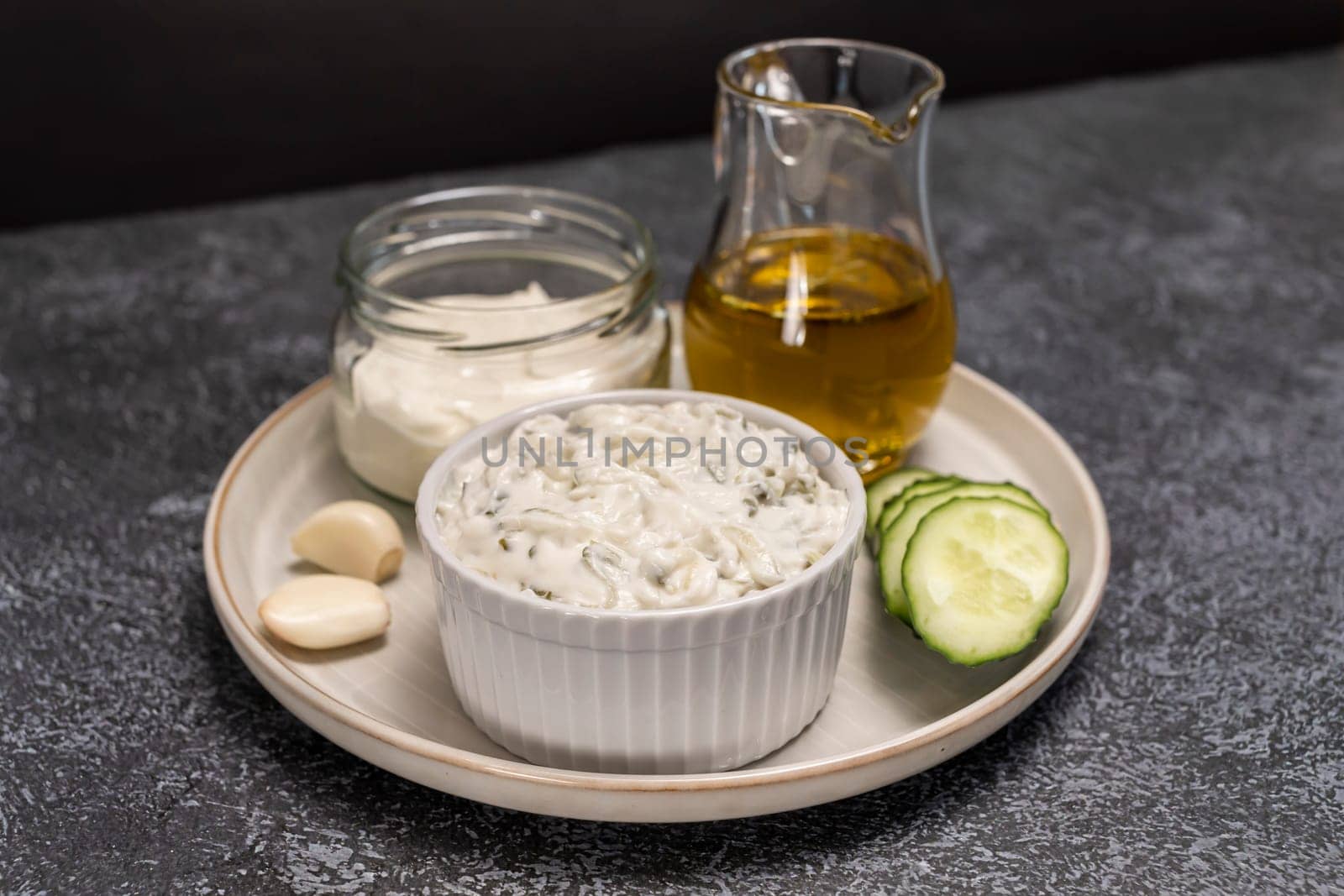 Traditional greek yogurt sauce Tzatziki in white bowl, made of chopped cucumbers, sour cream yogurt,olive oil,garlic,herbs with ingredients on on dark gray concrete table. Horizontal plane.