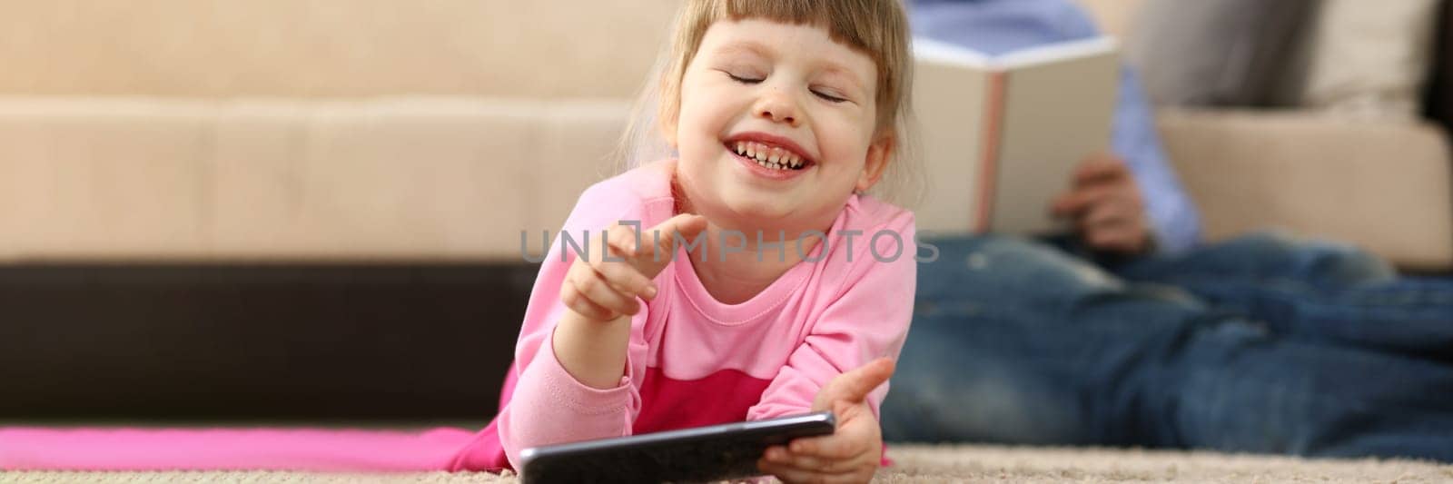 Little smiling baby girl with smartphone lies on floor in background dad reads a book. Children interests in phone and reading books