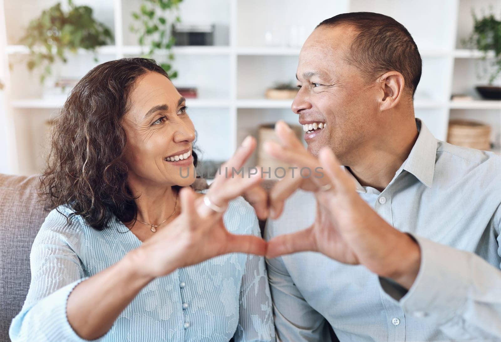 Hand, heart and mature couple with love on a sofa for bonding, romance and happy in a living room. Hands, emoji and romantic older man with woman, happy and smile while enjoying retirement lifestyle by YuriArcurs