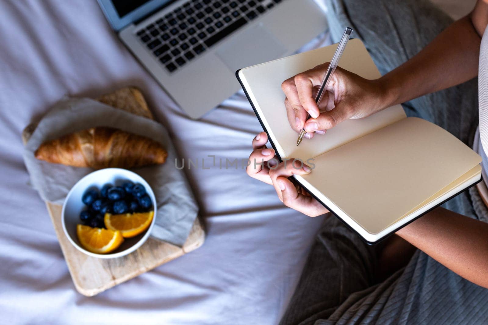 High angle view close-up of young unrecognisable woman hands writing on journal notebook sitting in bed writing having breakfast. Female working with laptop. Lifestyle concept.