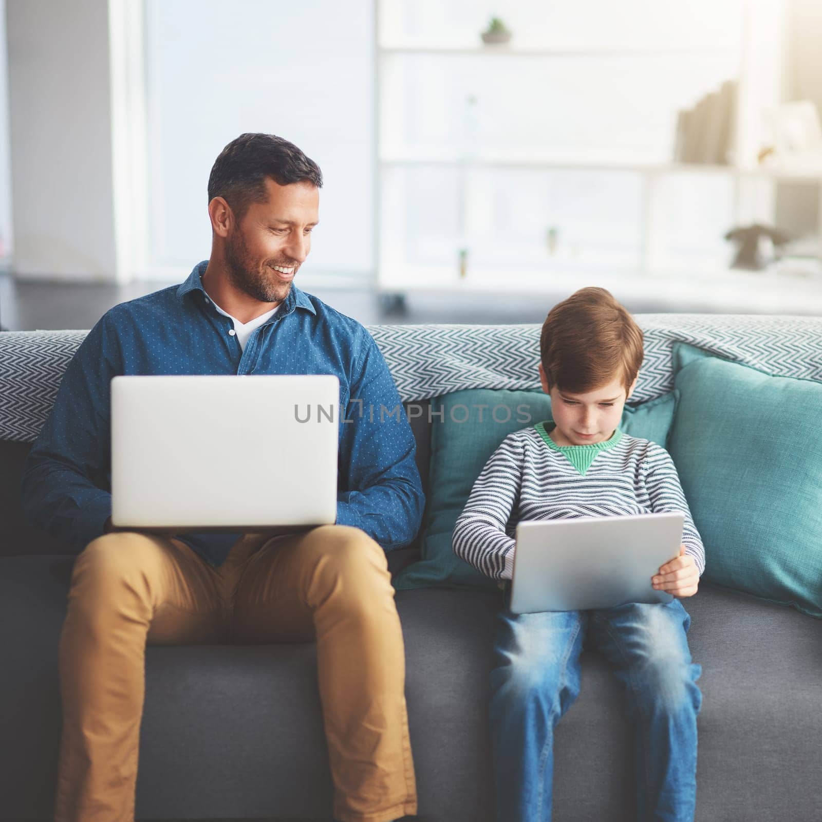 They are both serious about their work. a cheerful little boy and his father using a laptop and a digital tablet while being seated on a sofa at home during the day. by YuriArcurs