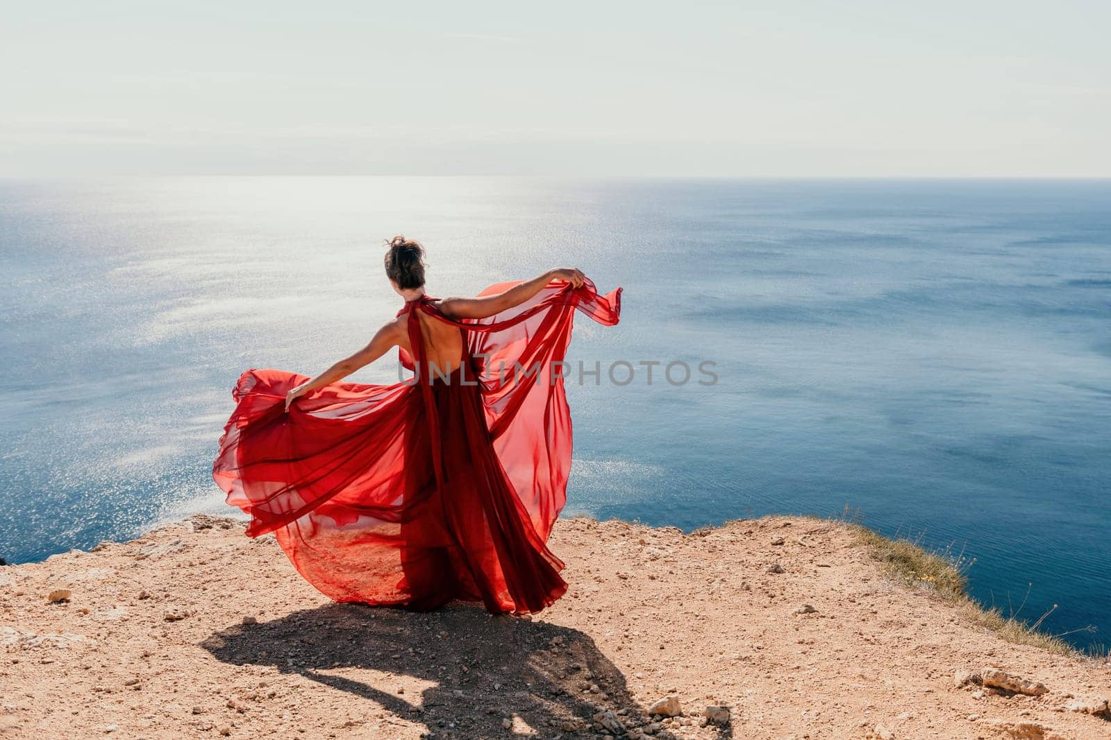 Side view a Young beautiful sensual woman in a red long dress posing on a rock high above the sea during sunrise. Girl on the nature on blue sky background. Fashion photo.