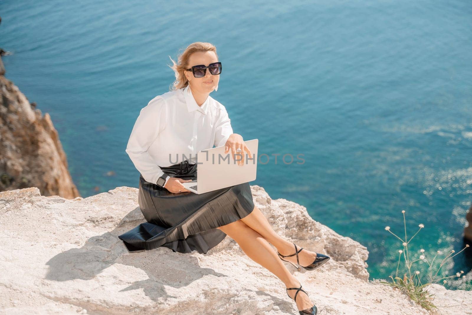 Business woman on nature in white shirt and black skirt. She works with an iPad in the open air with a beautiful view of the sea. The concept of remote work