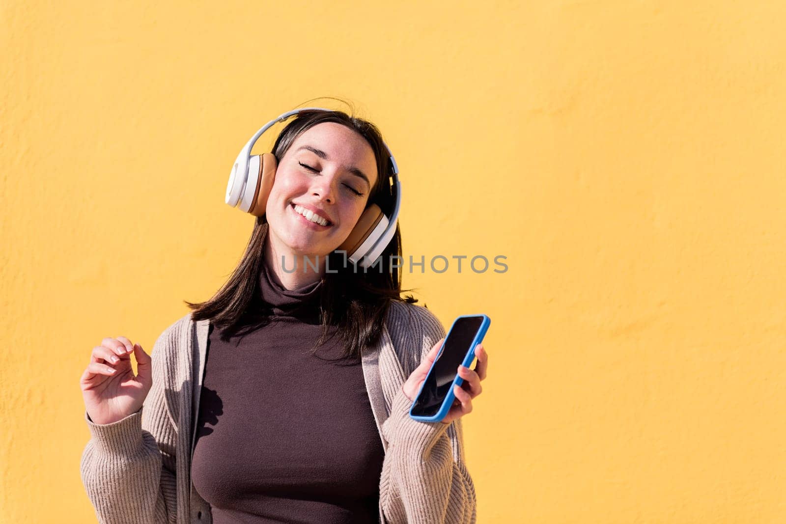 woman dancing happy listening to music from phone in headphones with a yellow wall in the background, concept of rhythm and positive people, copy space for text