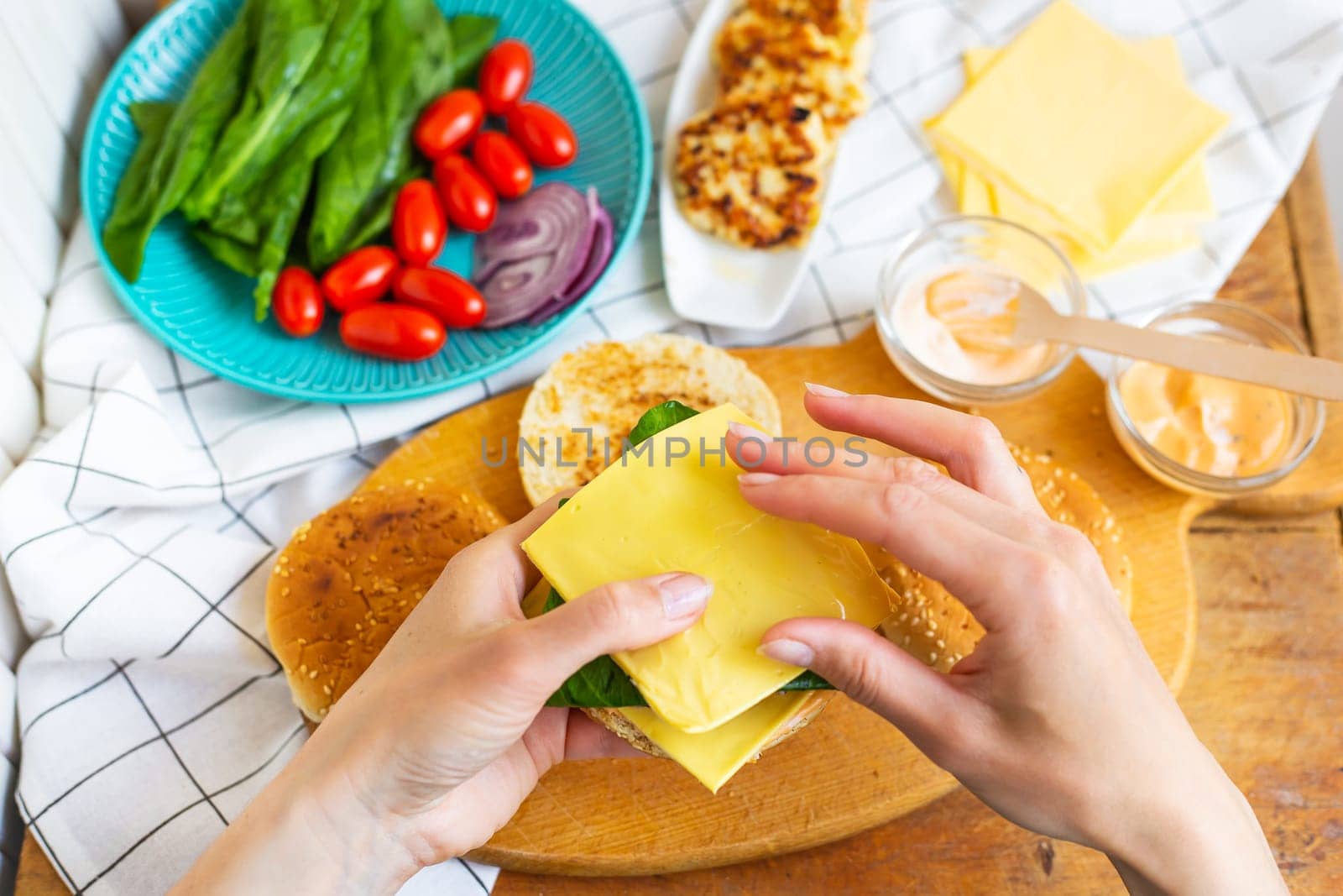 Preparation of all the ingredients for making a burger - bun, cutlet, cheese, salad, tomato, sauces. The girl s hands take a burger bun and put cheese on top of a cutlet