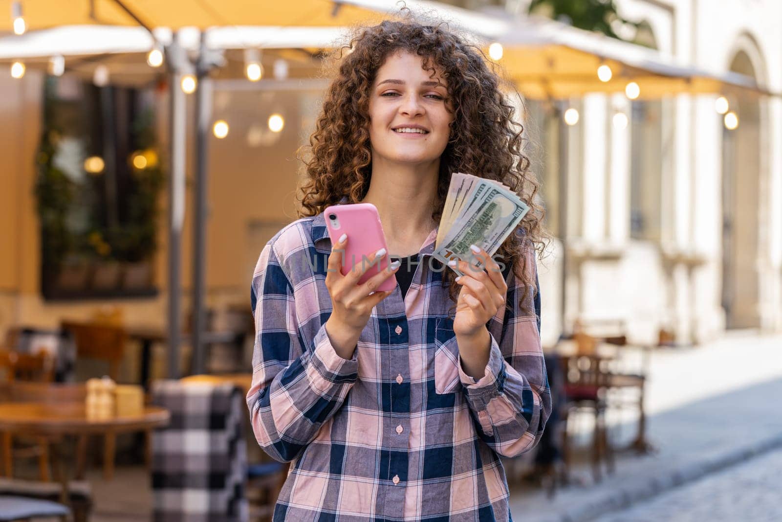 Happy rich young woman counting money dollar cash, use smartphone calculator app, plans to order gifts and food delivery online, booking hotel room. Pretty girl tourist in city street. Town outdoors