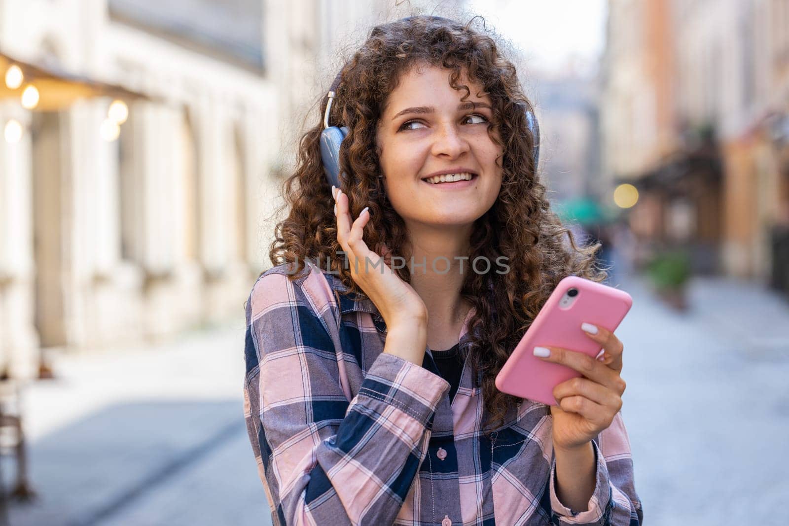Happy relaxed overjoyed pretty young woman in wireless headphones choosing, listening favorite energetic disco rock n roll music dancing outdoors. Girl walking in urban city sunshine street background