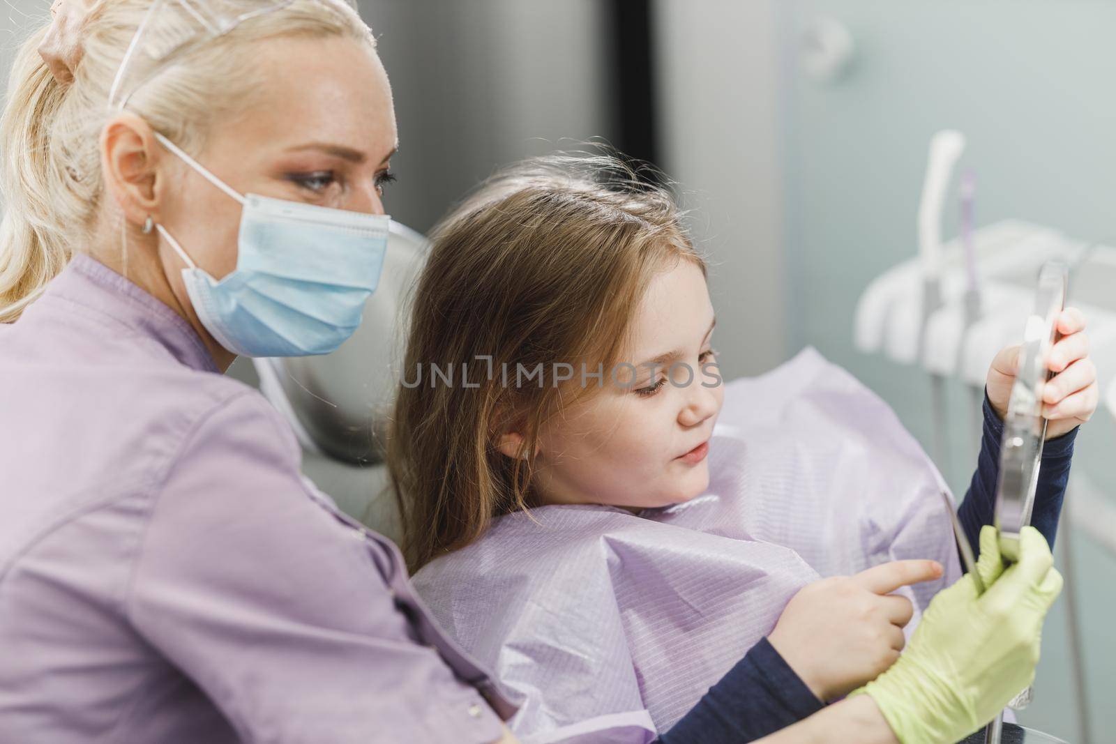 A female dentist and cute little girl's holding a mirror and looking her teeth after dental examination at dentist's office.