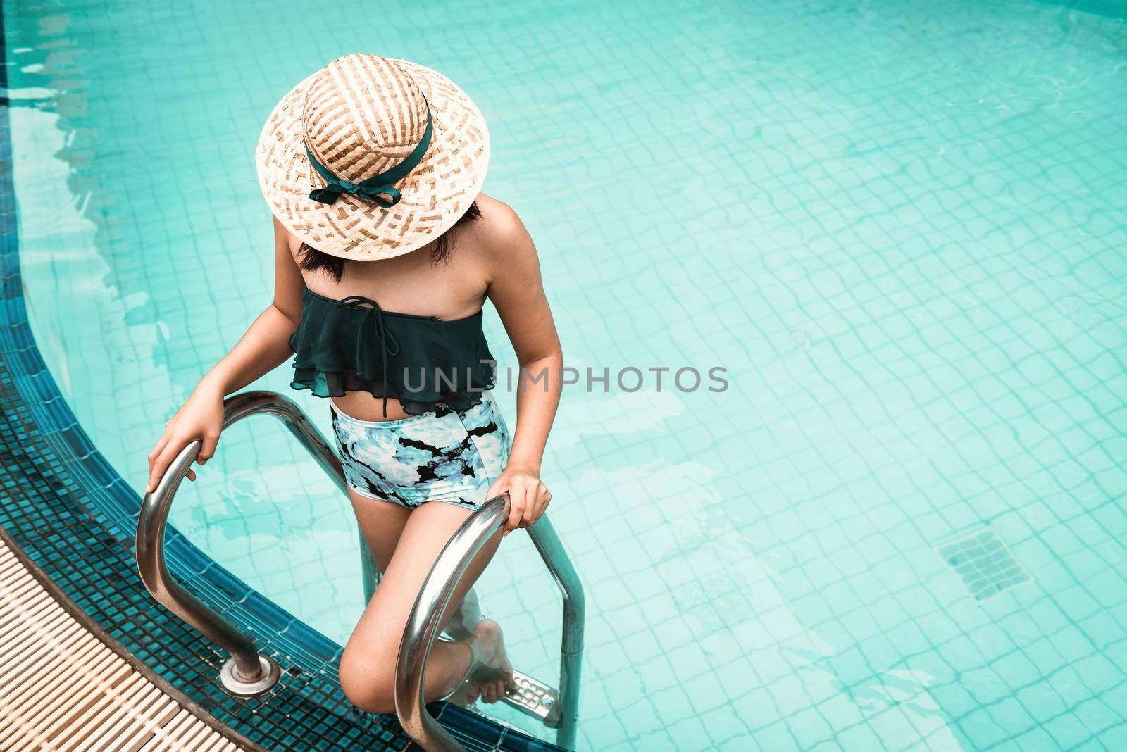 Sexy Woman in Swimsuit is Relaxing in Swimming Pool, Beautiful Woman Wearing Straw Hat and Relax in Poolside While Holding Pool Pedal Ladder at Resort Hotel. Summer Vacation and Relaxation Lifestyles by MahaHeang245789