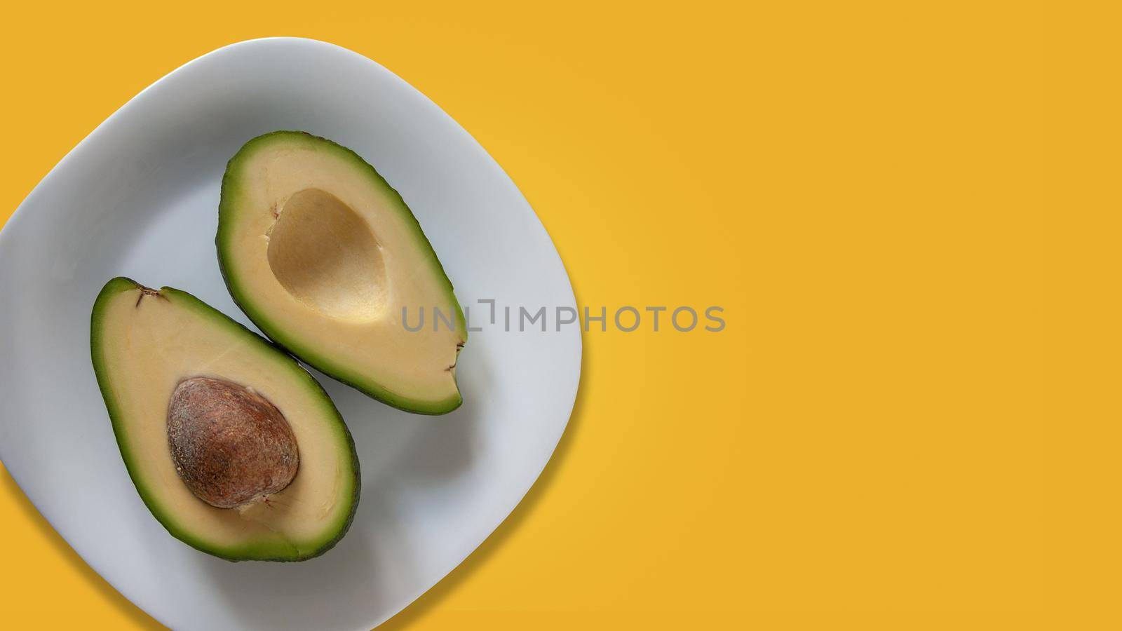 Two parts of avocado on white plate. Isolated on yellow background. 16x9 size. Template for banner with copy space at right side by LipikStockMedia