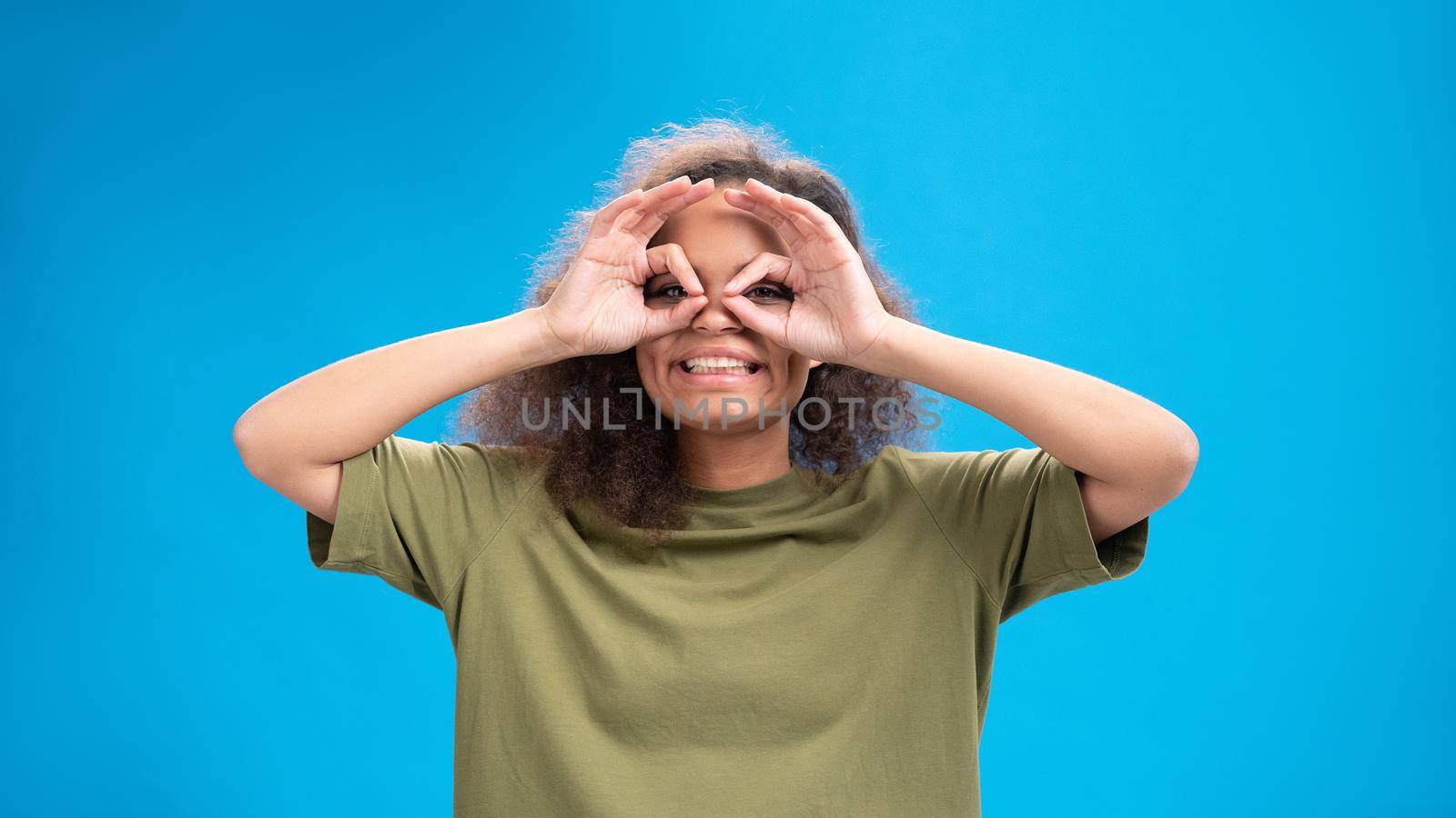 Funny pilot gesture African American girl looking positively at camera wearing olive t-shirt with hands as binoculars isolated on blue background. Beauty concept. 