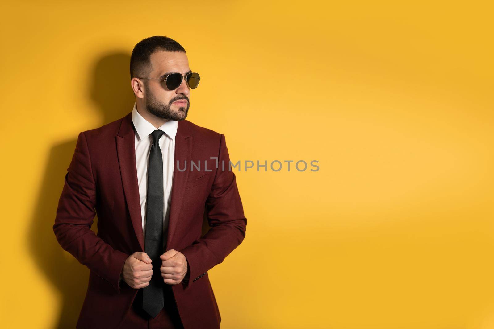 Young man in burgundy suit looking seriously sideways wearing sunglasses with hands holding a jacket isolated on yellow background. Men fashion concept by LipikStockMedia