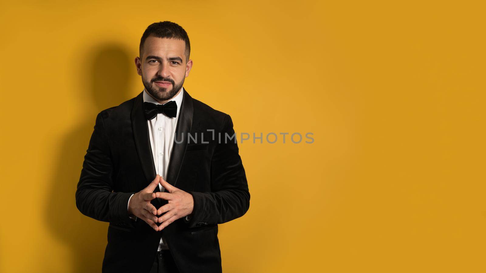 Handsome man in tuxedo standing fingers folded down smiling on camera, work profession lifestyle. Handsome young smiling caucasian man isolated on yellow background. 