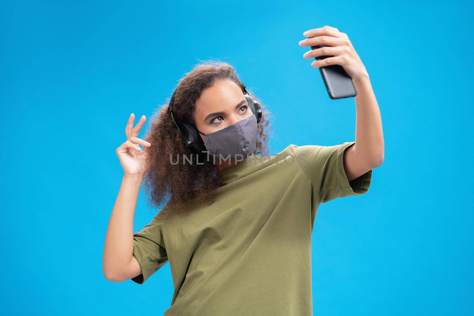 Dancing African American girl listen music on her smartphone with headphones in olive t-shirt, reusable face mask, to prevent others from corona COVID-19 infection isolated on blue background by LipikStockMedia