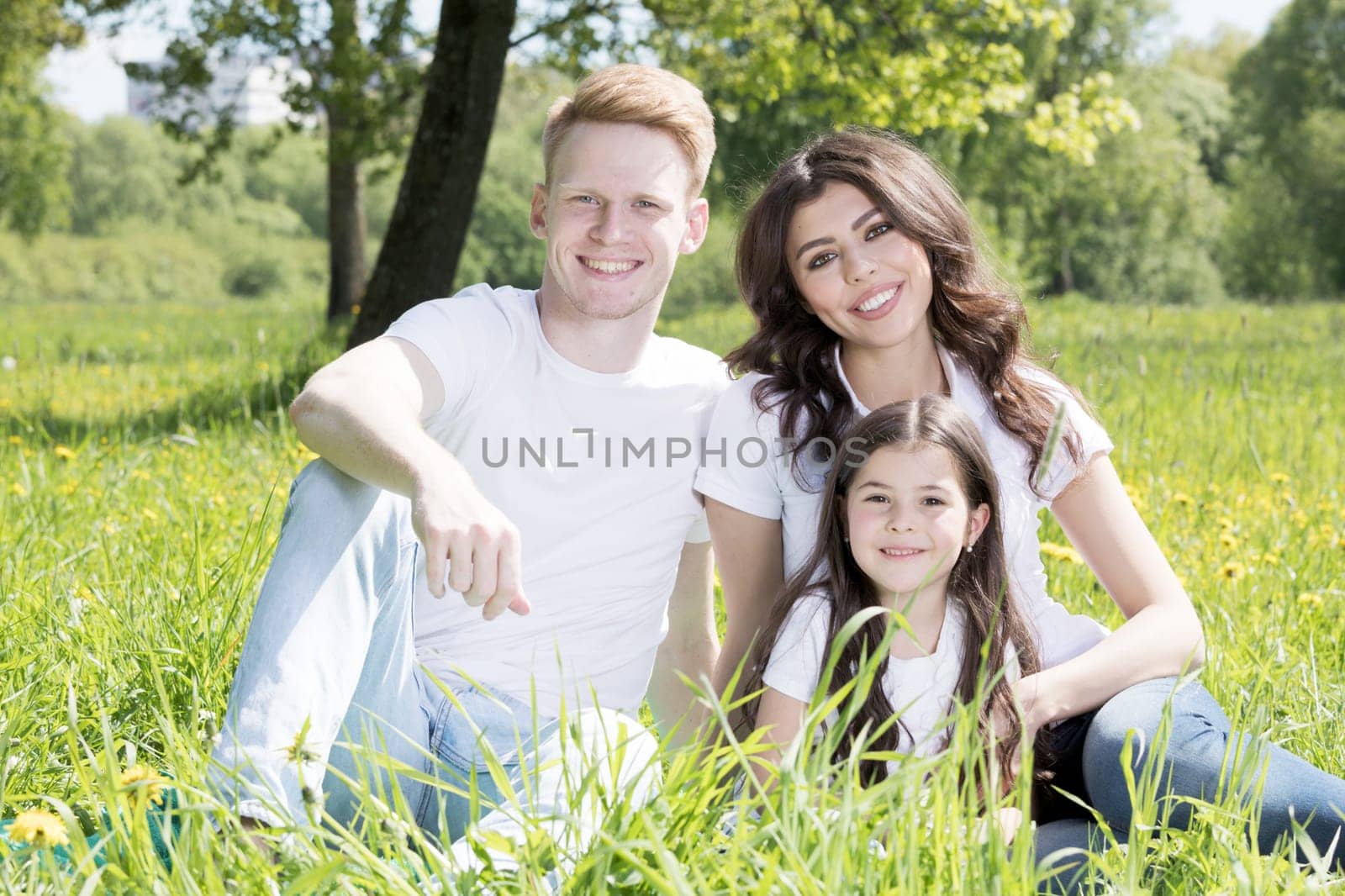 Portrait of happy smiling family of parents and girl sitting on grass with dandelion flowers at sunny summer day