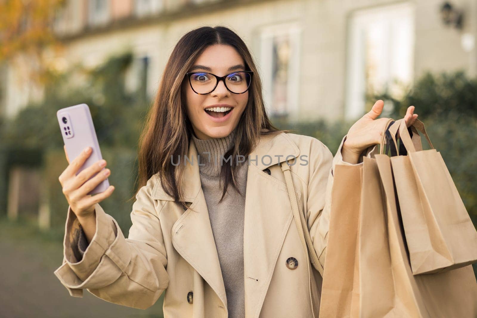 Portrait of attractive happily surprised woman with glasses holding eco-carton shopping bags and a phone in urban street.