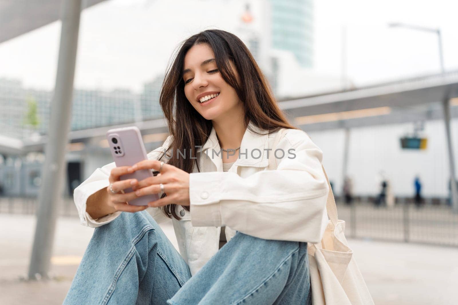 Bright and cheerful young woman with shopper on shoulder waiting public transport at station smiling beautifully while texting on phone.
