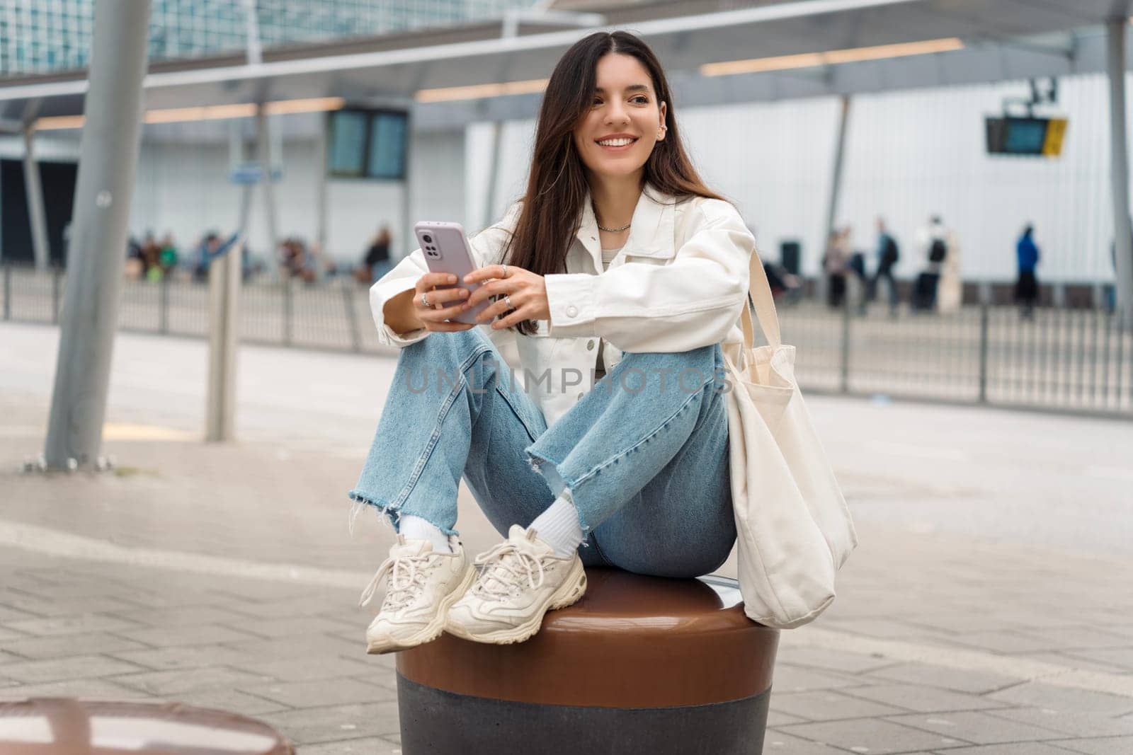 Bright portrait of cheerful young woman 25s with shopper on shoulder waiting public transport at station smiling beautifully and texting by AndreiDavid