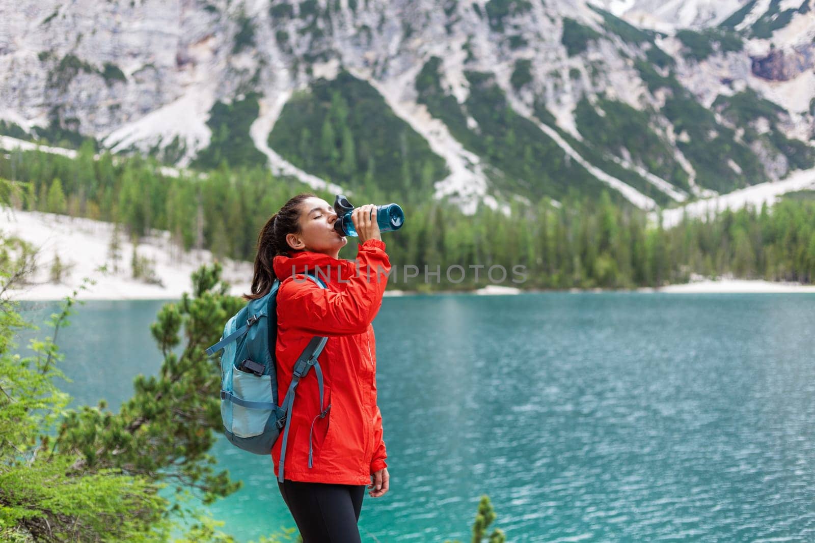 Hydration while on track. Attractive woman hiker in red raincoat drinking water near a lake with mountains in the background.