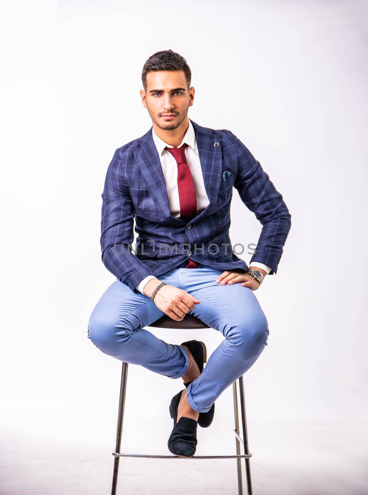 Handsome young man in blue business jacket and jeans posing on white background in studio, sitting on stool. Full body shot. Looking at camera
