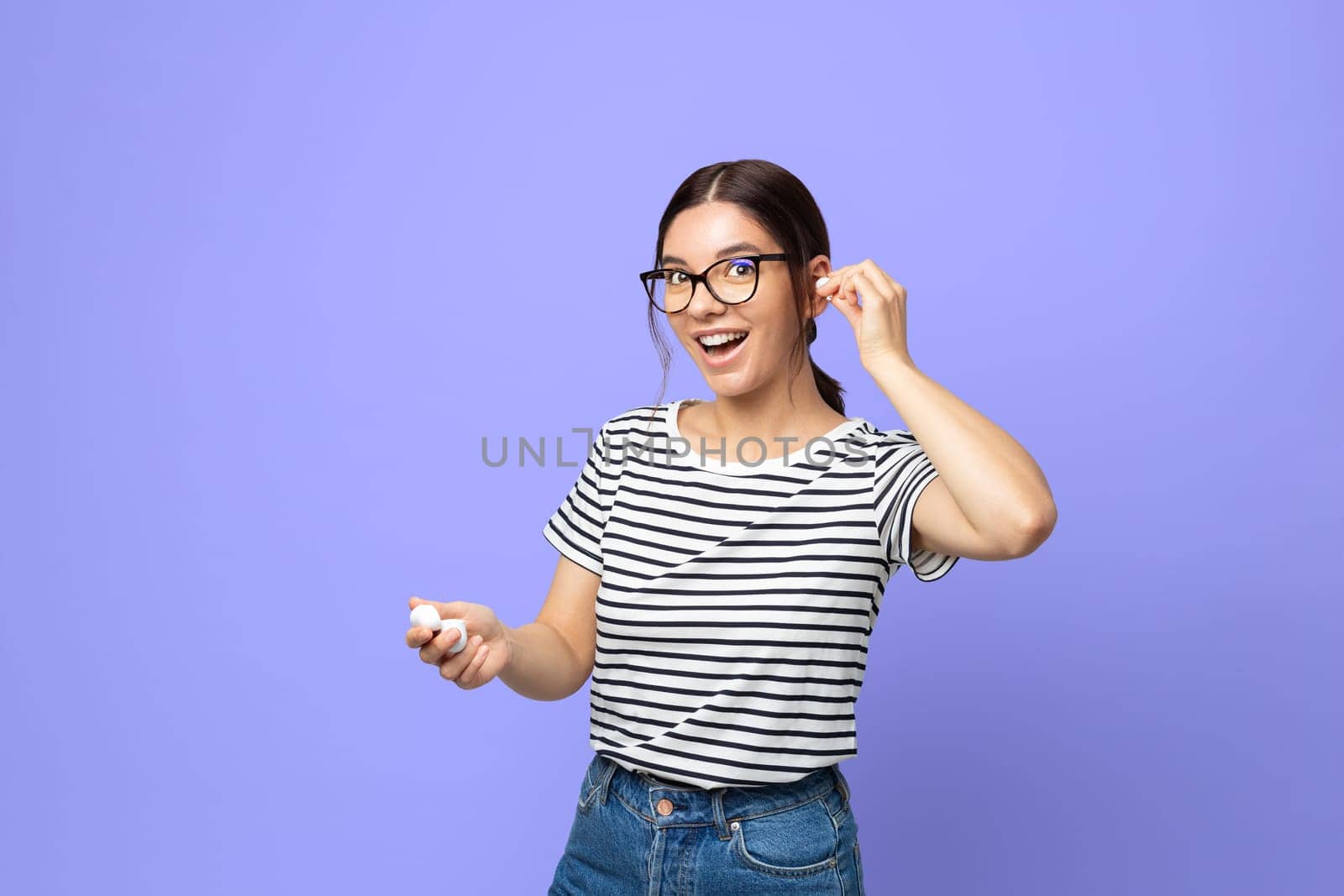 attractive brunette in striped t-shirt using small white wireless headphones against purple very peri color background.