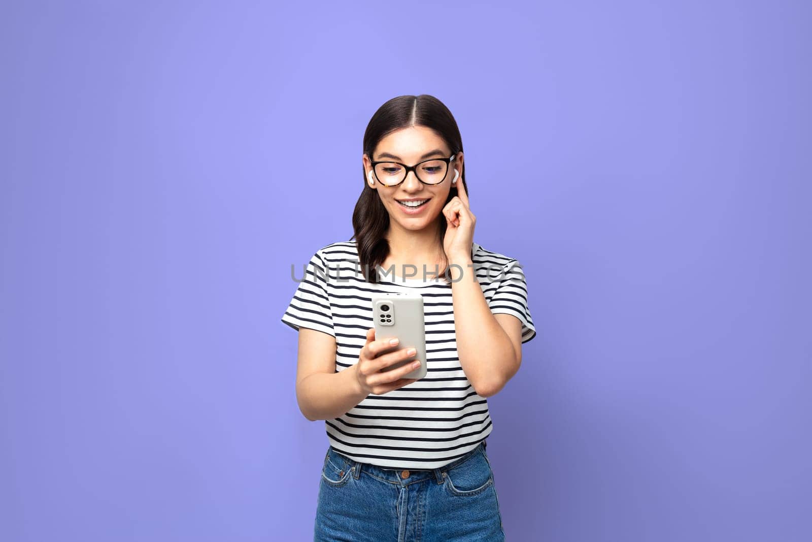 attractive brunette in striped t-shirt using small white wireless headphones against purple very peri color background.