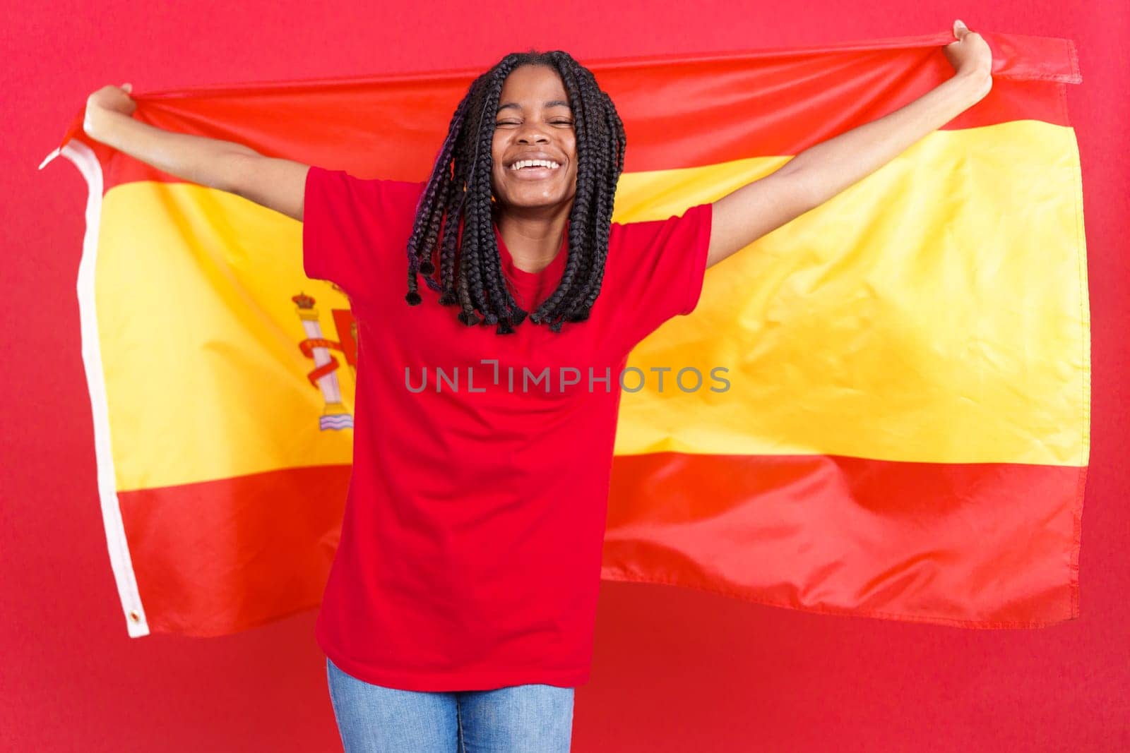 Happy african woman smiling and raising a spanish national flag in studio with red background