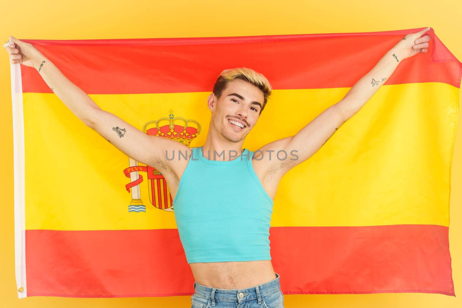 Happy gay man smiling and raising a spanish national flag in studio with yellow background