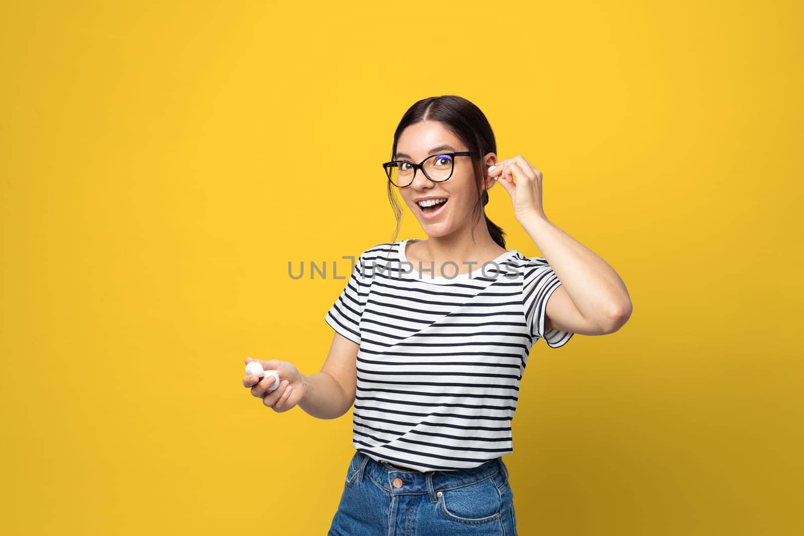 Funny attractive multi ethnicity young woman with glasses using in-ears wireless headphones against yellow background.