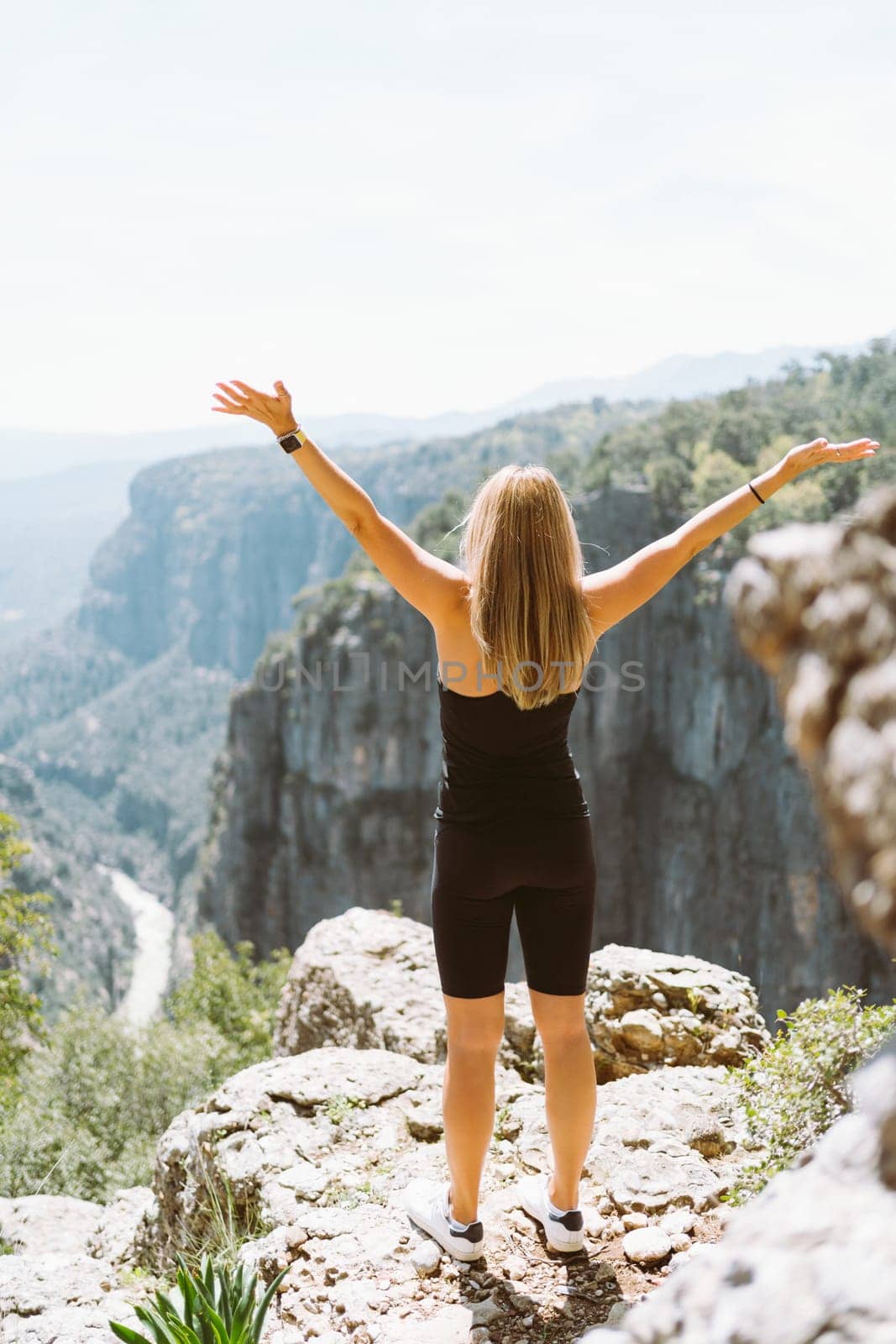 Young fit beautiful girl woman in black sporty slim outfit standing on edge of mountain cliff peak with hands up in air enjoying view of canyon. Picture back female hiker reached the mountain top by Ostanina