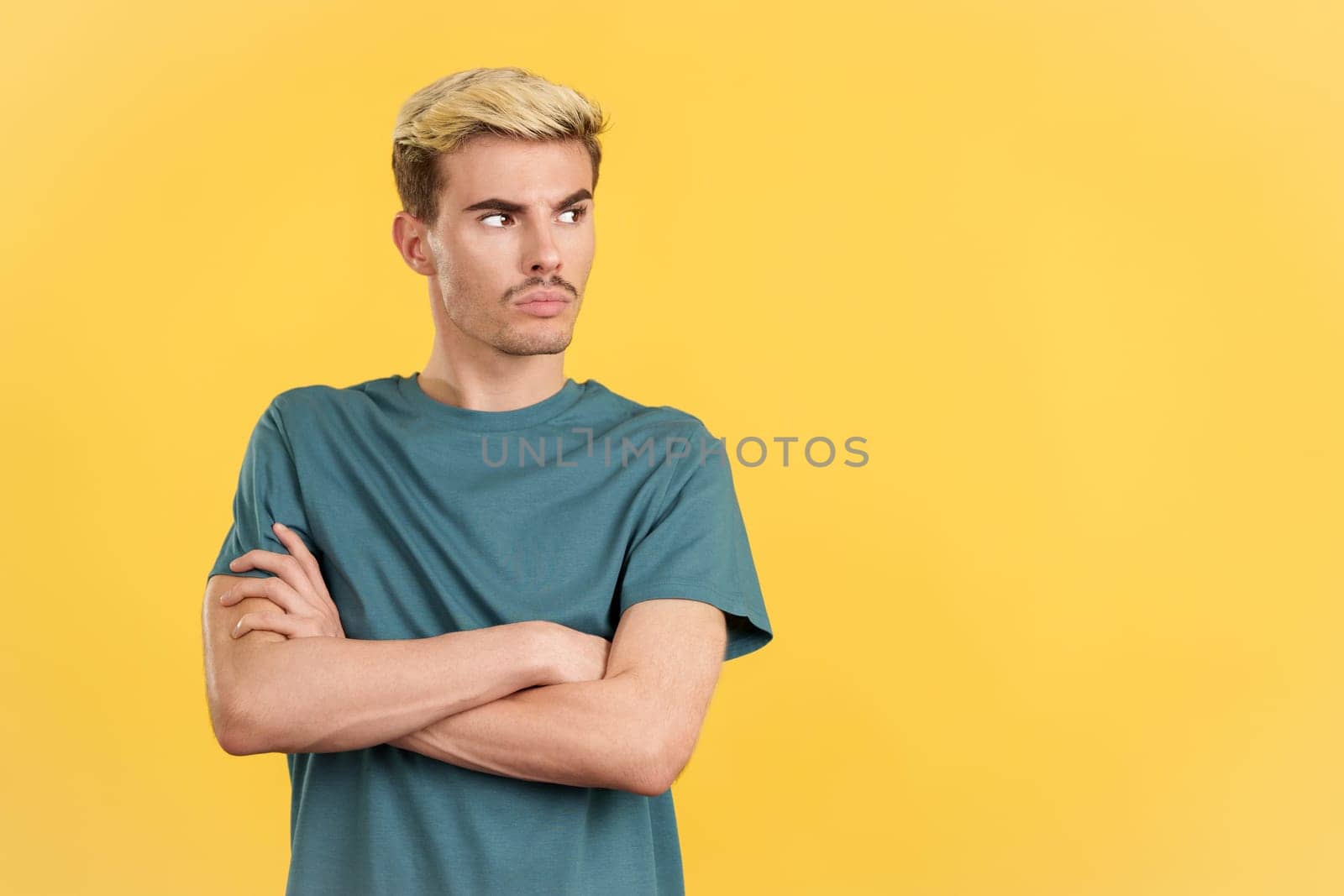 Angry gay man standing looking away with arms crossed in studio with yellow background