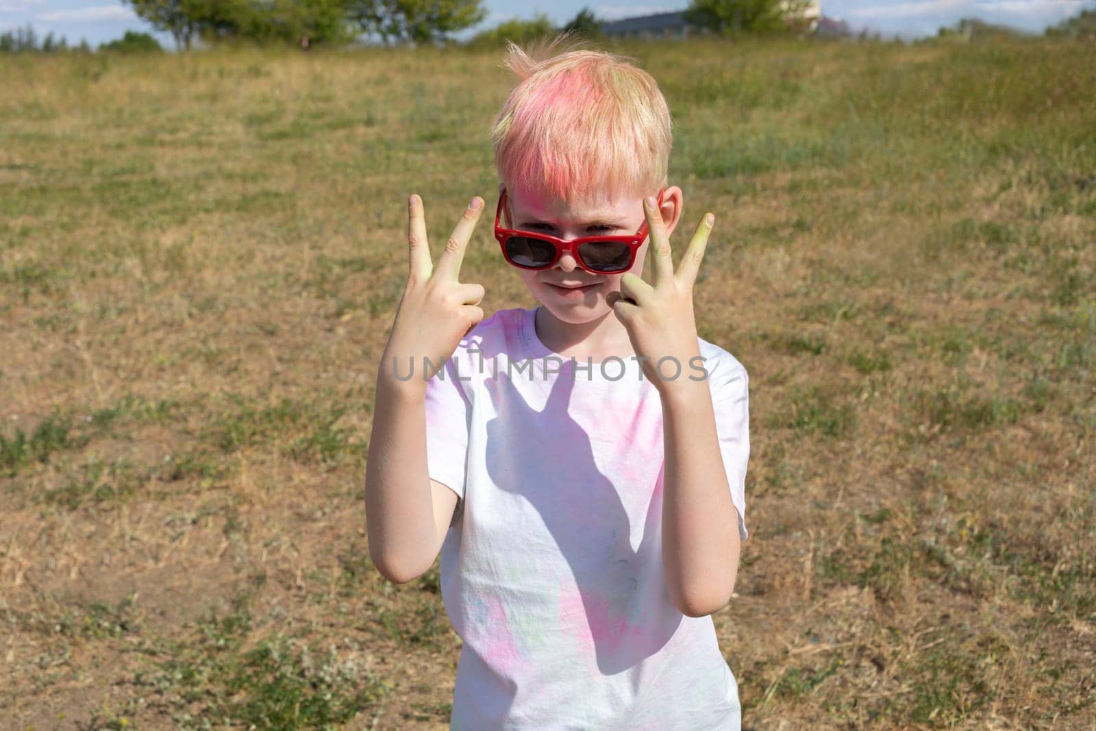 Little Blonde Male Child With Colorful Paint, Powder On Clothes Posing On Holi Color Festival In Meadow. Emotional Happy Child. Blue Sky, Green Trees On Background, Sunny Day. Horizontal Plane,