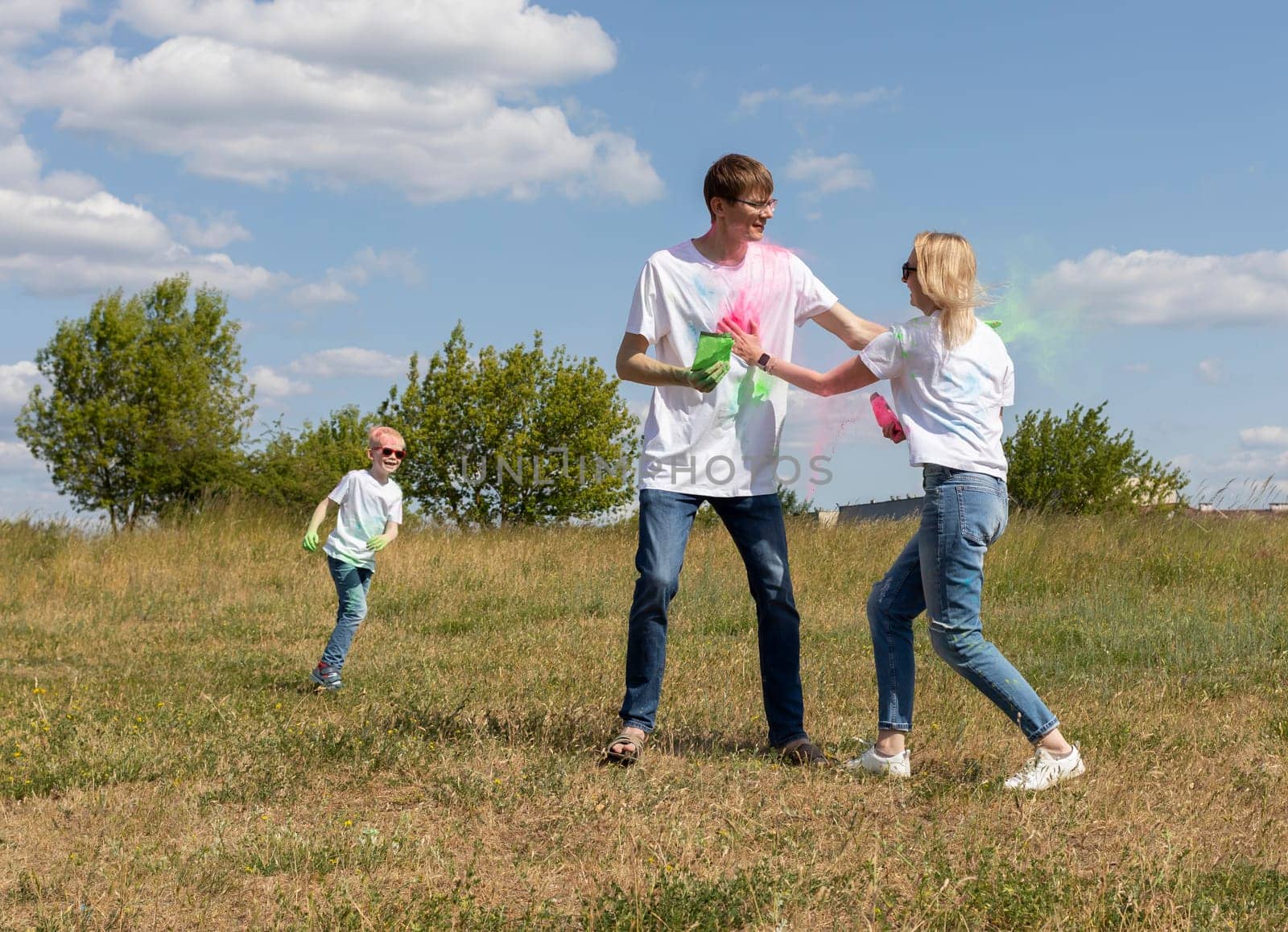 Caucasian Family Plays With Colorful Dye At Birthday Party Or Celebrates Holi Color Festival, Throwing Multicolor Powder. Cheerful Parent, Child Plays With Paint Outdoor In Park. Horizontal Plane.