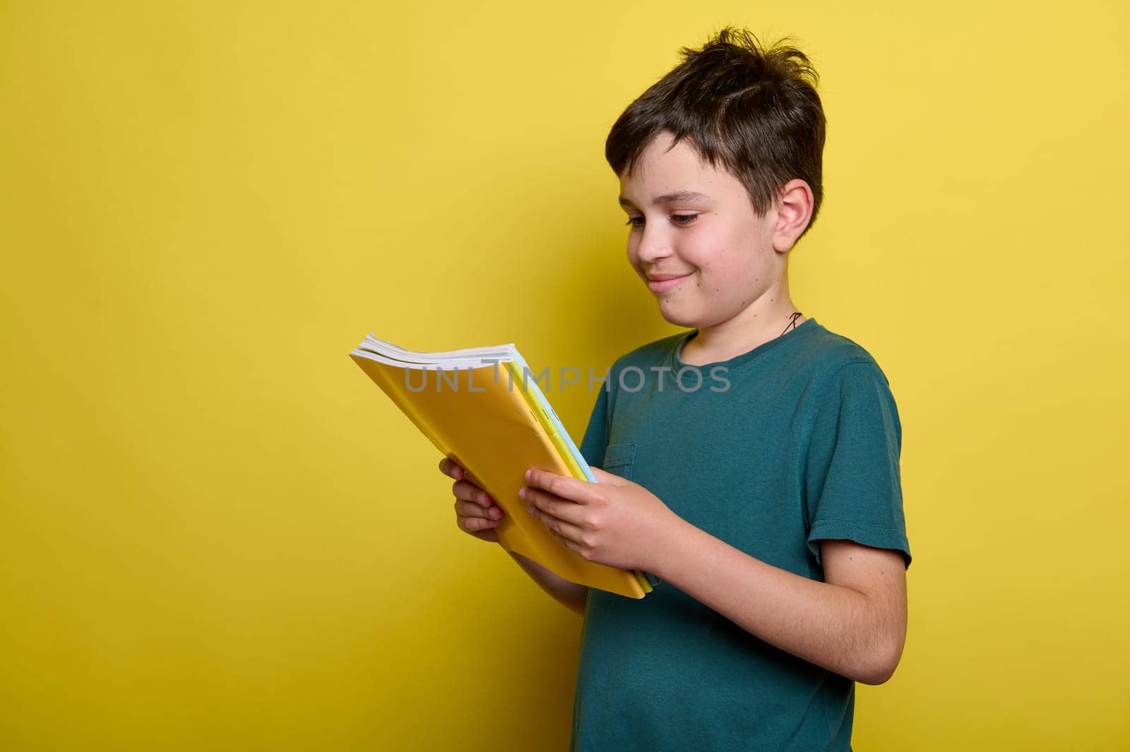 Caucasian adorable teenager school boy 10-13 years old, smart pupil, holding books, smiling standing isolated on yellow background. Copy advertising space. Education. Erudition. Back to school