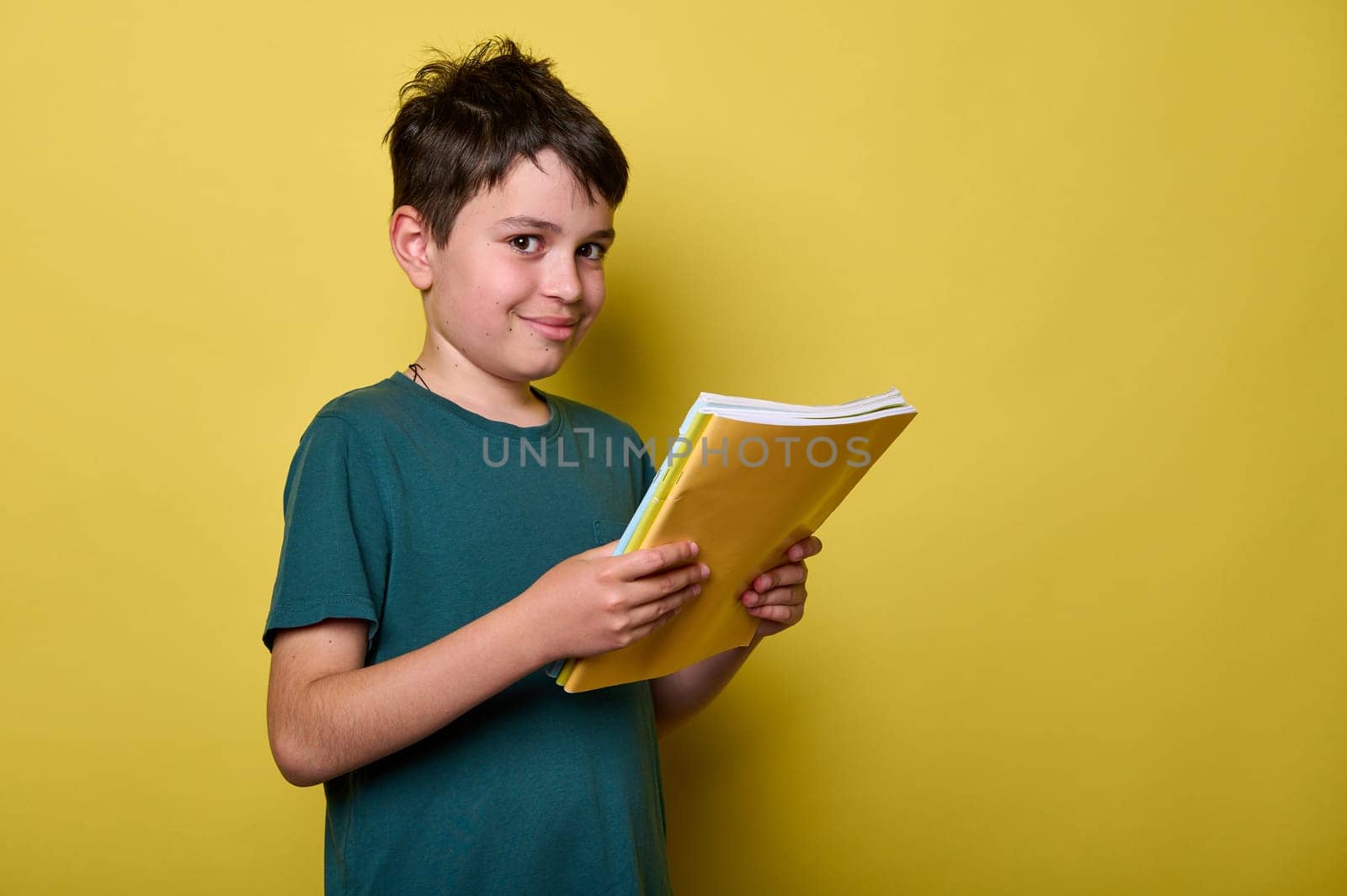 Handsome Caucasian teenage boy 10-12 years old in green casual t-shirt, smiling looking at camera, holding textbooks, ready to start a new semester of academic school year, isolated yellow background