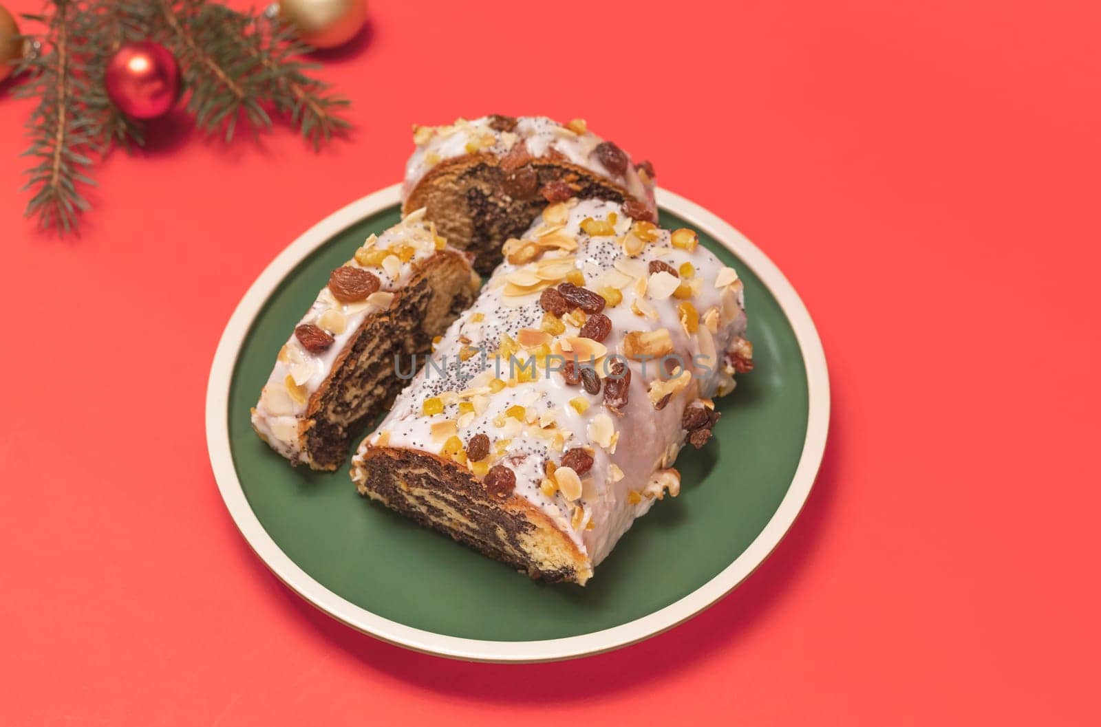Christmas strudel with poppy seeds, traditional polish festive pie Makowiec on green plate,covered with sugar icing,walnuts,dried fruits on red background with Christmas tree, toy balls. Horizontal.