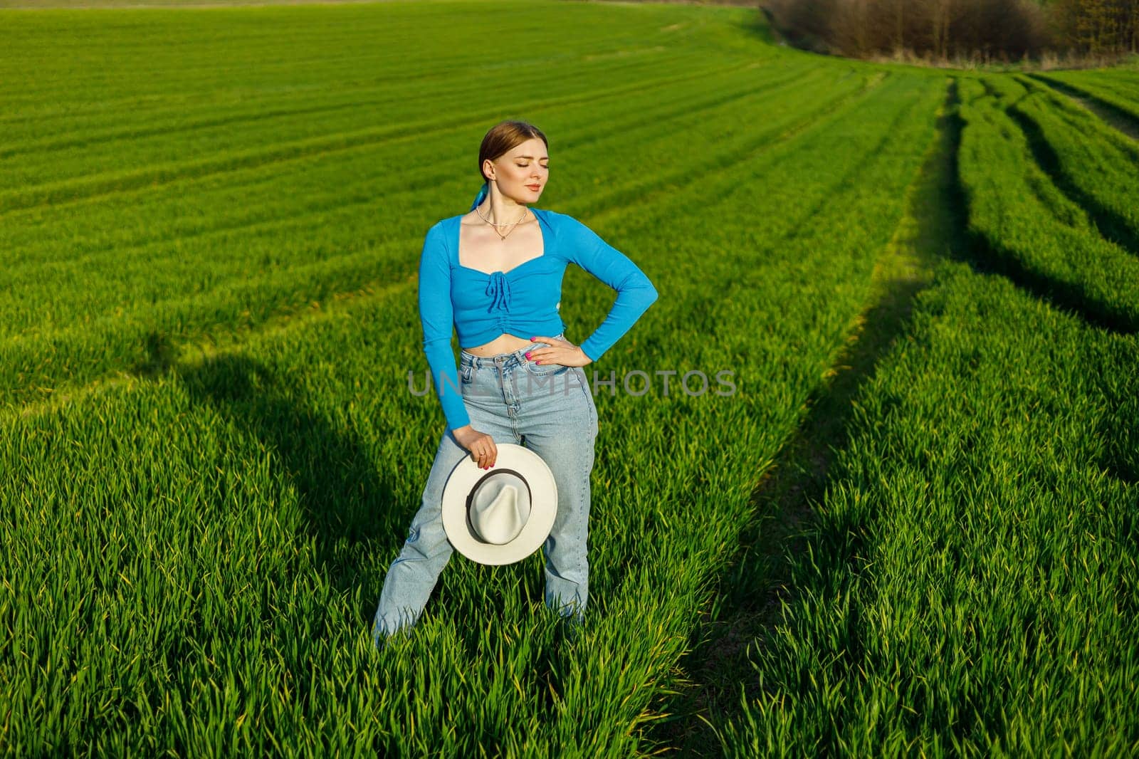 A cute woman in jeans and a hat stands in a green field. A smiling woman in a blue top and jeans walks in the green grass.