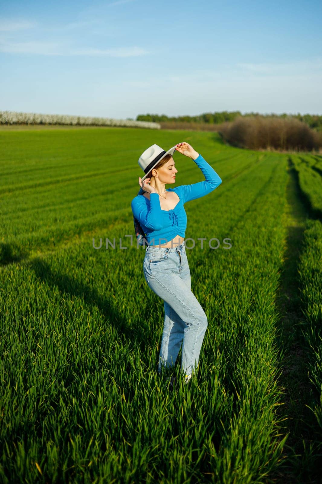 A cute woman in jeans and a hat stands in a green field. A smiling woman in a blue top and jeans walks in the green grass.